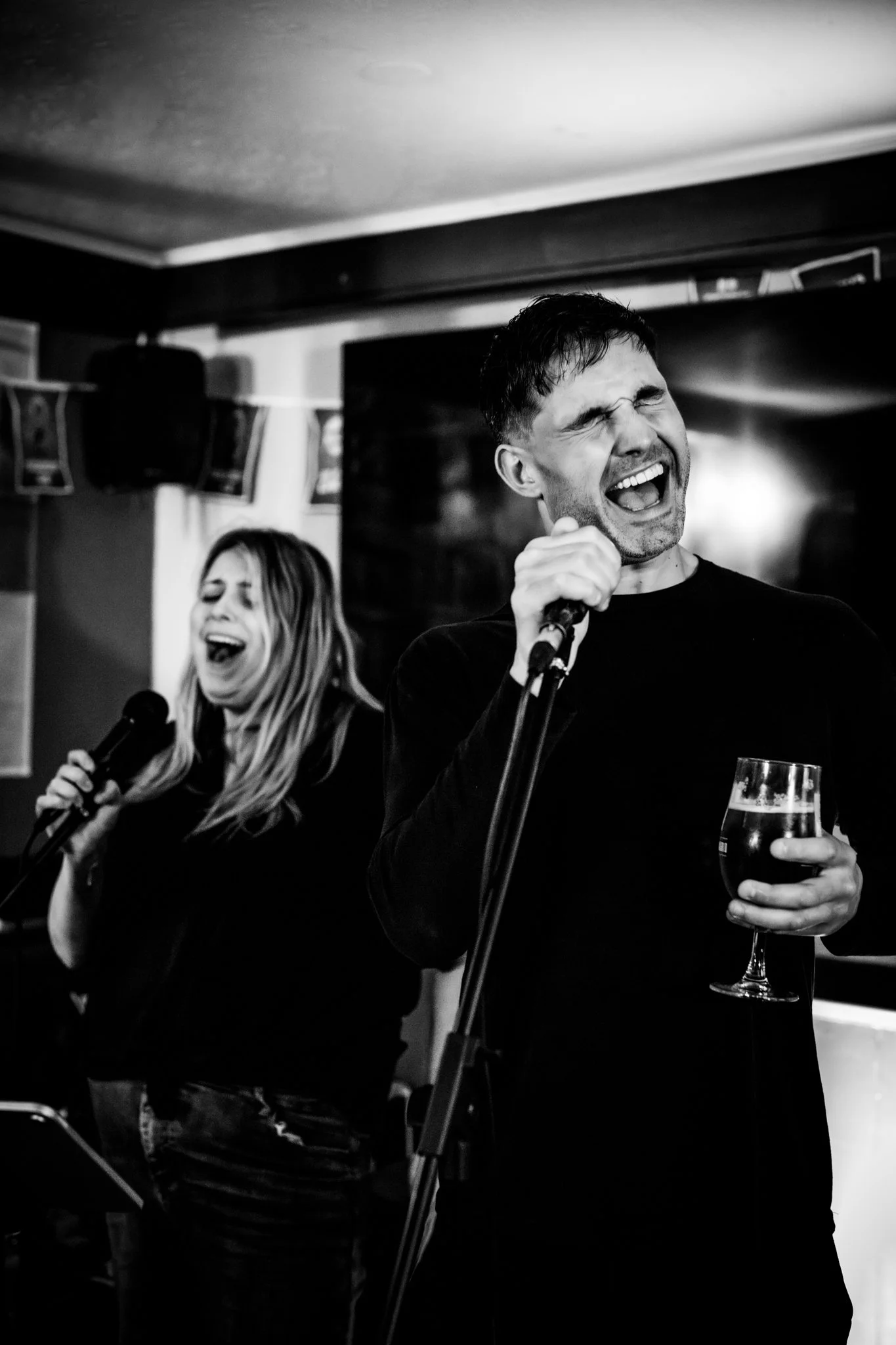 A man and a woman singing with microphones in a bar or pub. The man is holding a glass of wine and appears to be singing passionately with his eyes closed and mouth wide open. The woman is also singing, holding the microphone with one hand, and has h