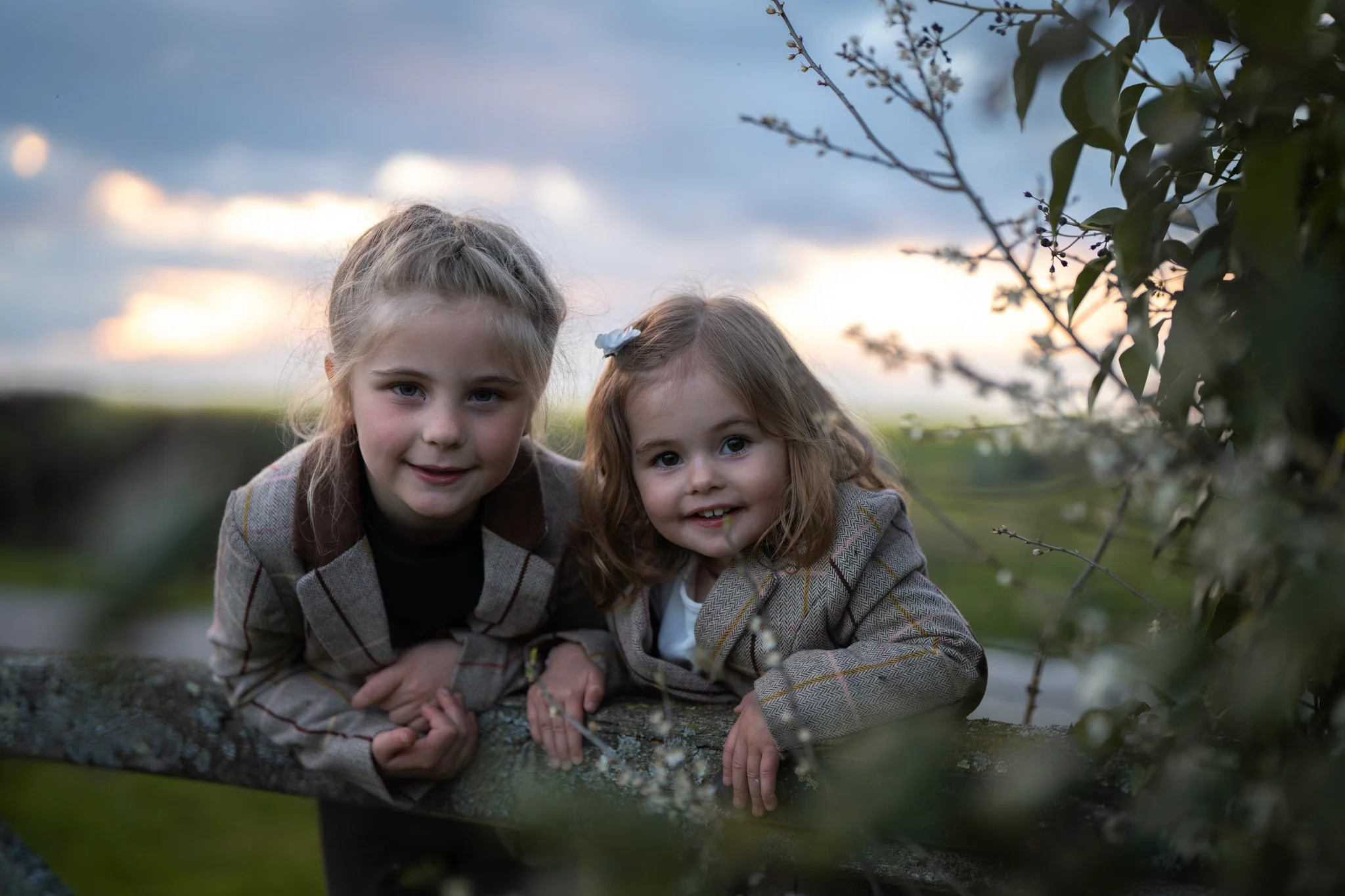 Two young girls with blonde hair, wearing plaid jackets, lying on a large log outdoors during dusk with cloudy sky and branches of a tree nearby. feasted, Essex. Anne Wills Photography 