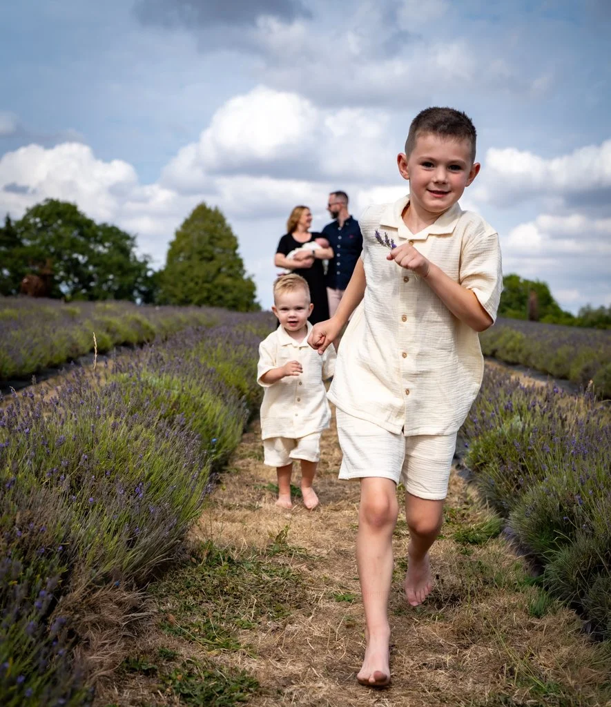 Two young boys running barefoot through a lavender field, with a happy couple holding a baby in the background under a cloudy sky.