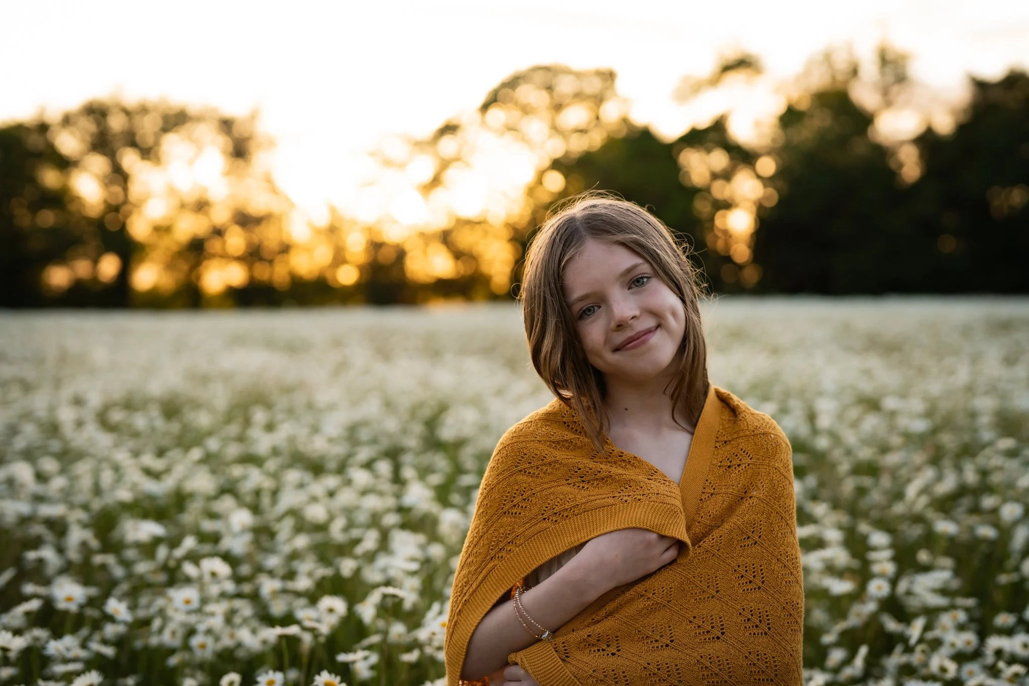 A young woman with light brown hair and wearing a mustard yellow, crocheted shawl, standing in a field of white daisies at sunset, smiling gently at the camera.