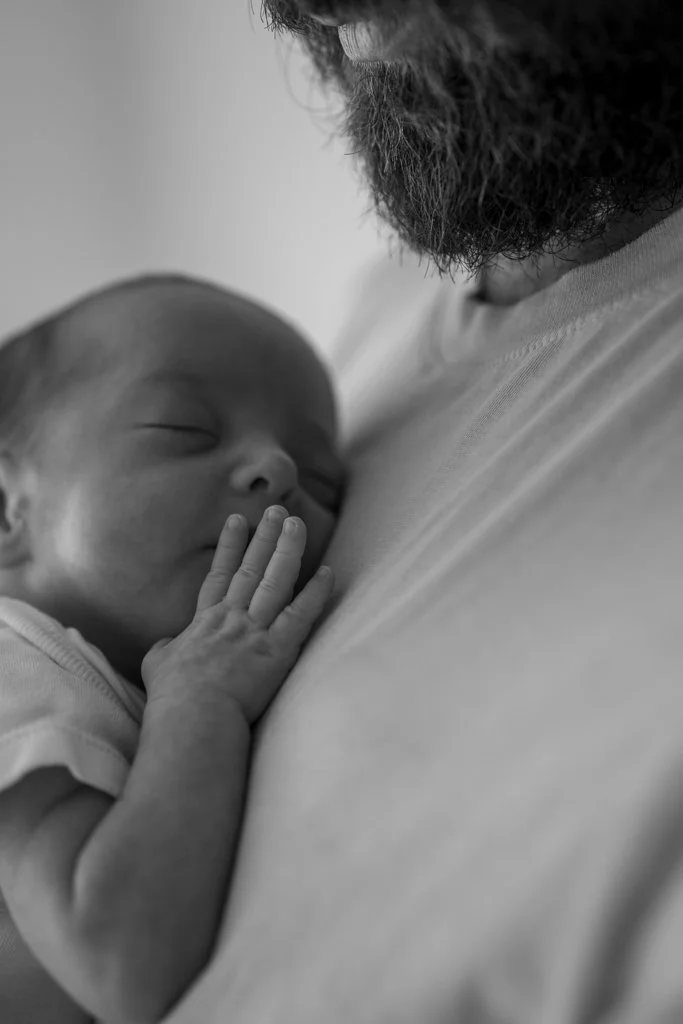 Close-up black and white photo of a sleeping baby resting on an adult's chest, with the adult's beard and a portion of their face visible.