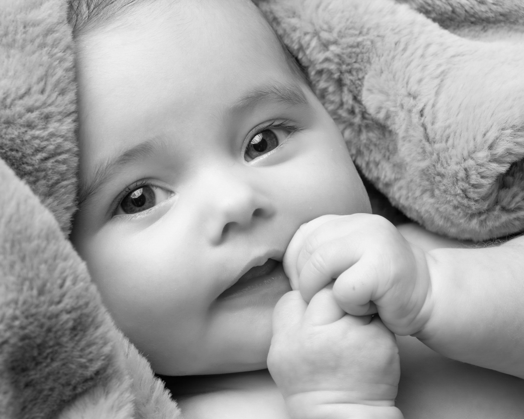 Close-up black and white photo of a young child with wide eyes, lying on a soft, plush blanket, holding a pacifier.