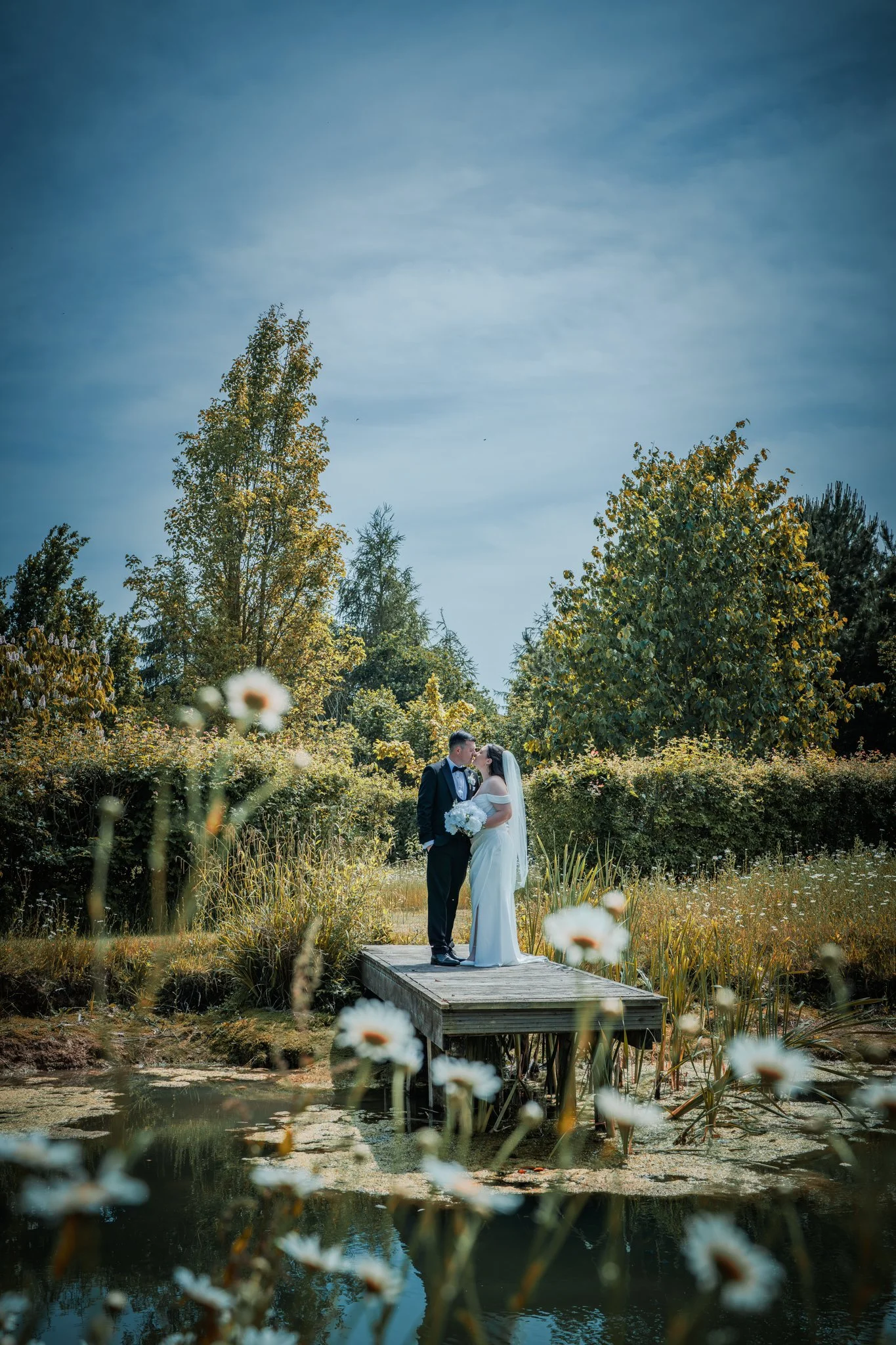 A bride and groom standing on a wooden dock by a pond, kissing, with trees and bushes in the background under a blue sky. Anne Wills Photography Maidens barn Essex