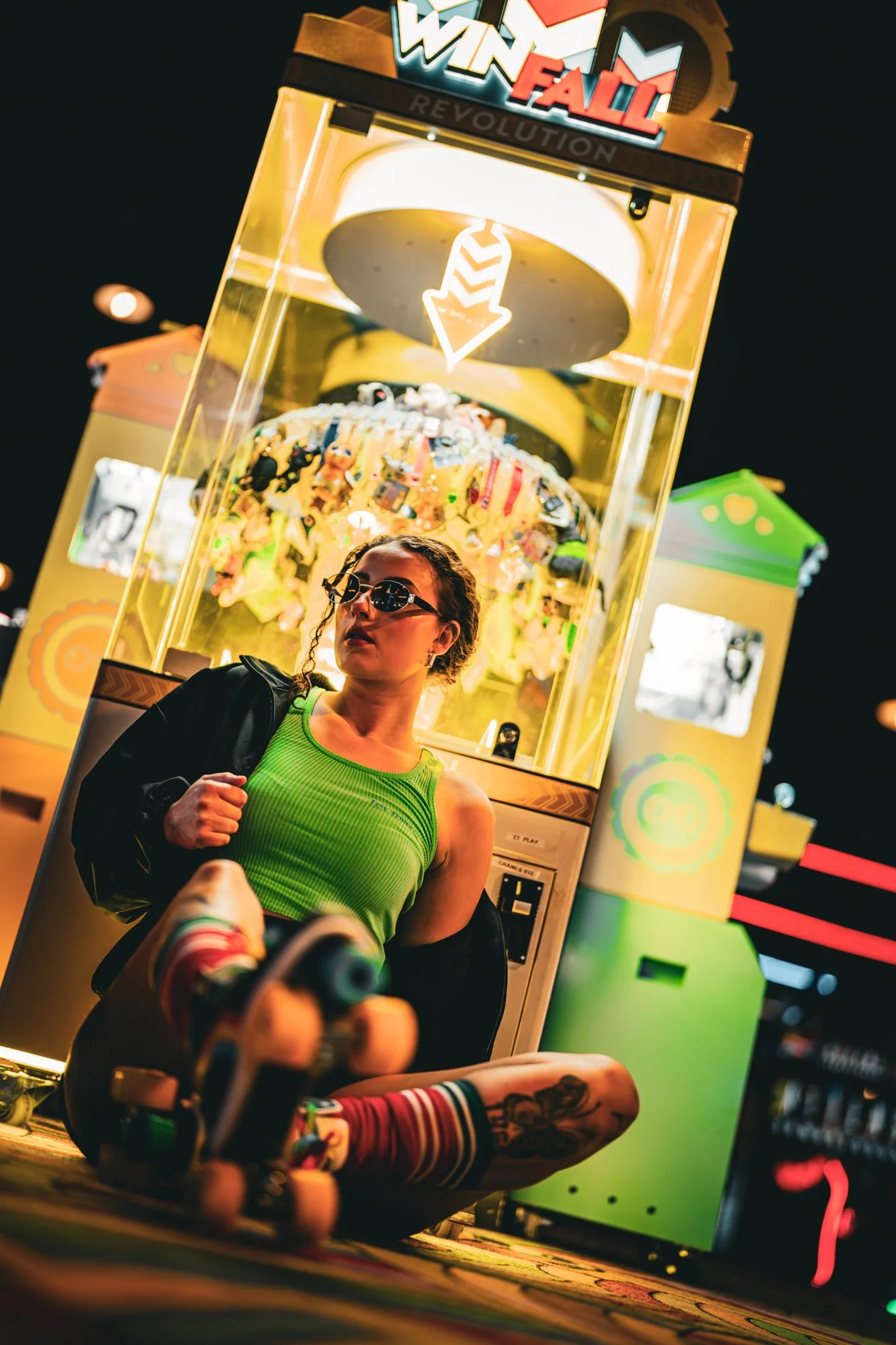 A woman wearing sunglasses and a green tank top sitting on the floor at a night fair, next to a claw machine game with plush toys. She has a black jacket over her shoulder and colorful striped socks. photographed in Walton, essex in the arcades on th