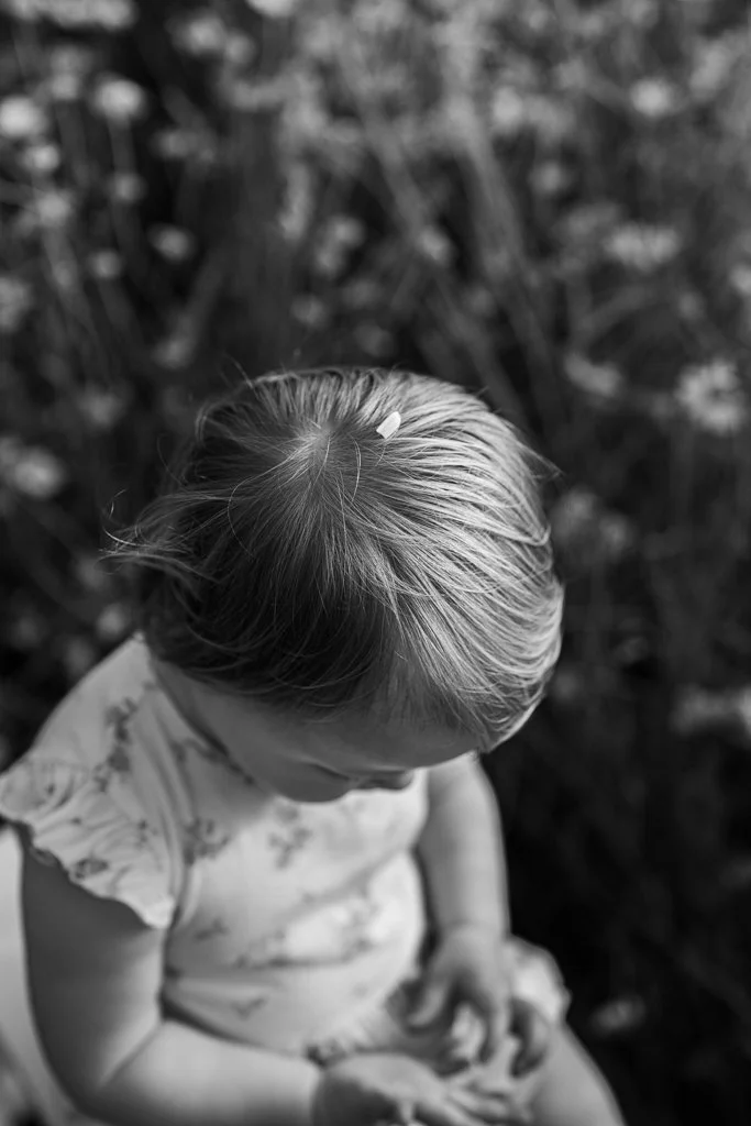A young girl with light-colored hair and a floral dress, seen from above, sitting outdoors with blurred foliage in the background. Anne Wills Photography. Braintree, Essex