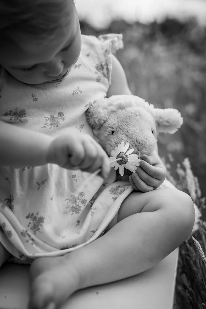A young child sitting outdoors on a blanket, holding a plush toy and a flower. Anne Wills Photography. Braintree, Essex
