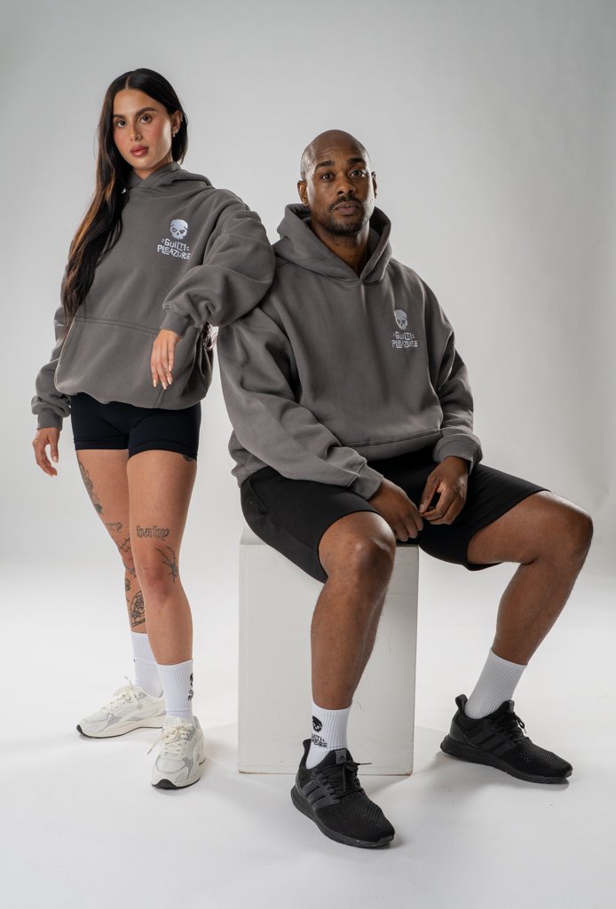 A young woman and a man wearing gray hoodies with a skull logo and text, black shorts, and athletic shoes pose against a plain white background; she stands with her right hand on her thigh, and he sits on a white cube, looking at the camera.