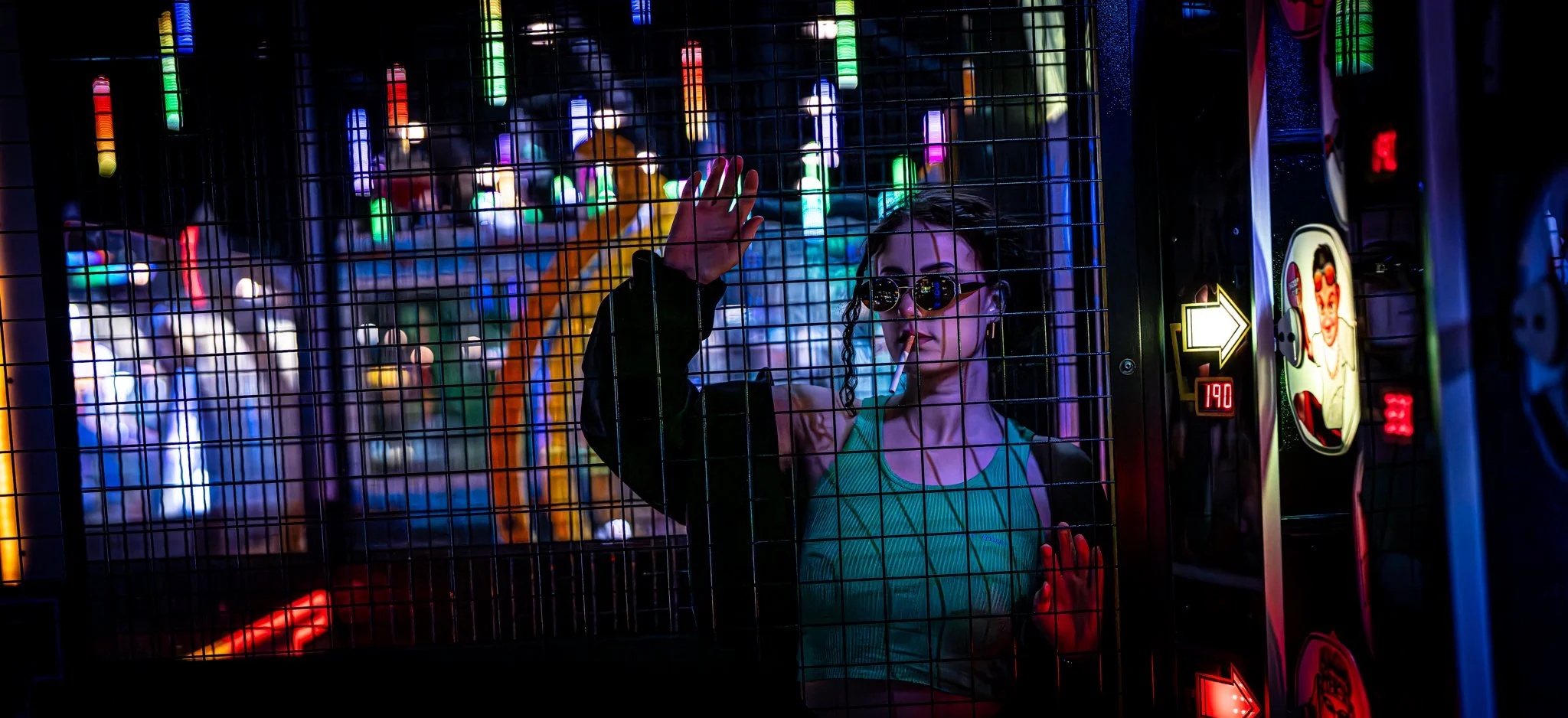 A woman with sunglasses is playing a basketball arcade game, seen through a metal grid at an amusement center with colorful neon lights in the background.