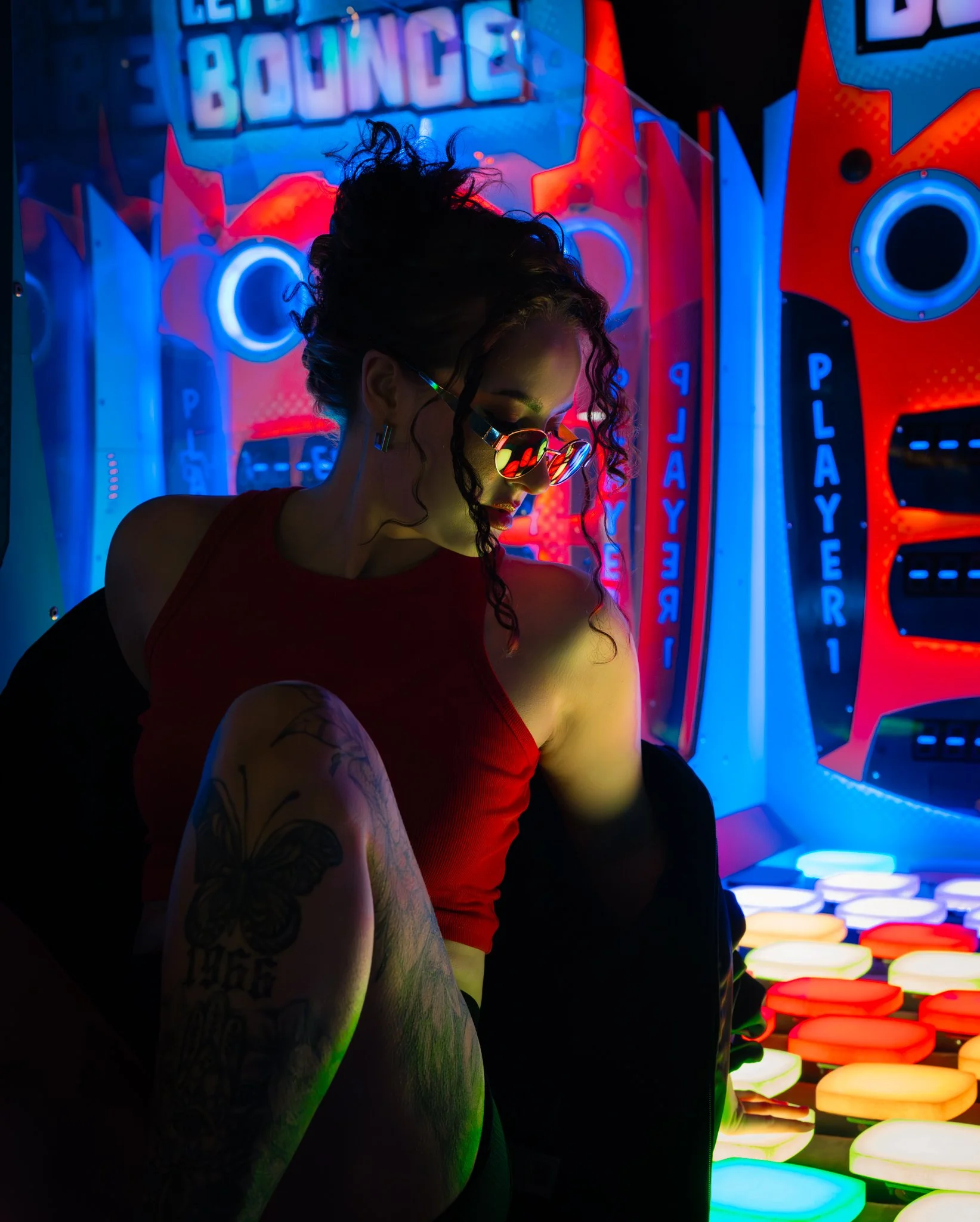 A woman with curly hair, sunglasses, and tattoos on her left arm stands in front of a colorful neon-lit arcade game. The background includes illuminated signs with words like "RELAY" and "BOUNCE" and rows of glowing, round, multicolored seats. photog