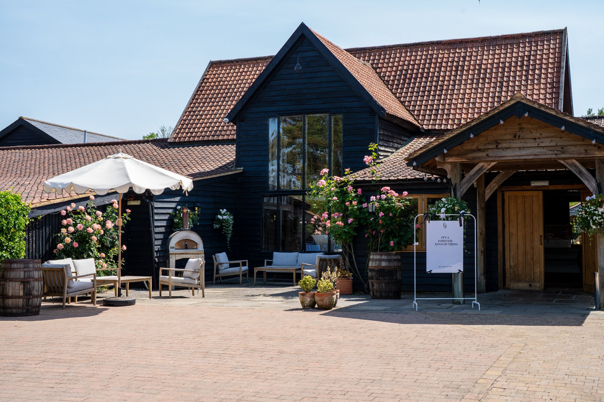 An outdoor patio area in front of a dark-colored wooden building with a sloped tiled roof. There are chairs, potted plants, and a white umbrella, and the sky is clear. Anne Wills Photography Maidens barn Essex