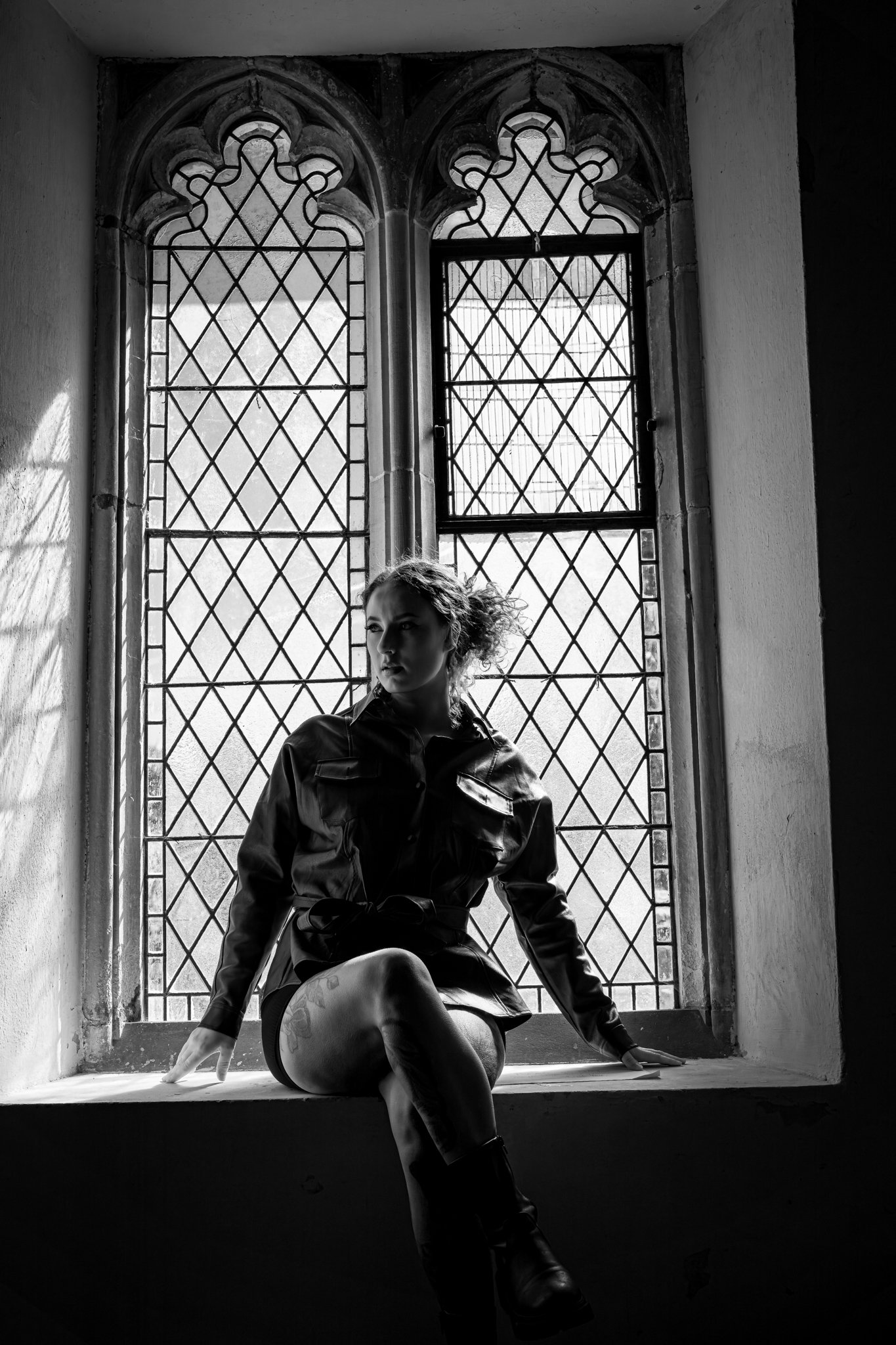 A woman with curly hair sitting on a window ledge in front of an ornate, arched stained glass window, with sunlight casting shadows on the wall behind her.