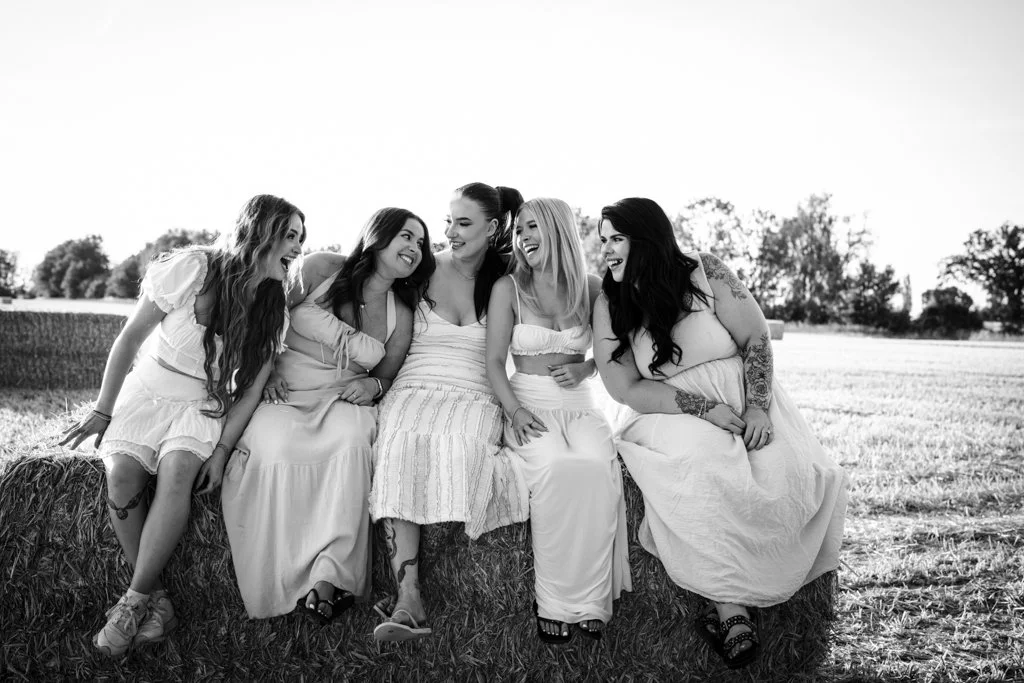 Five women wearing white dresses sitting on hay bales in a field, smiling and laughing together outdoors.