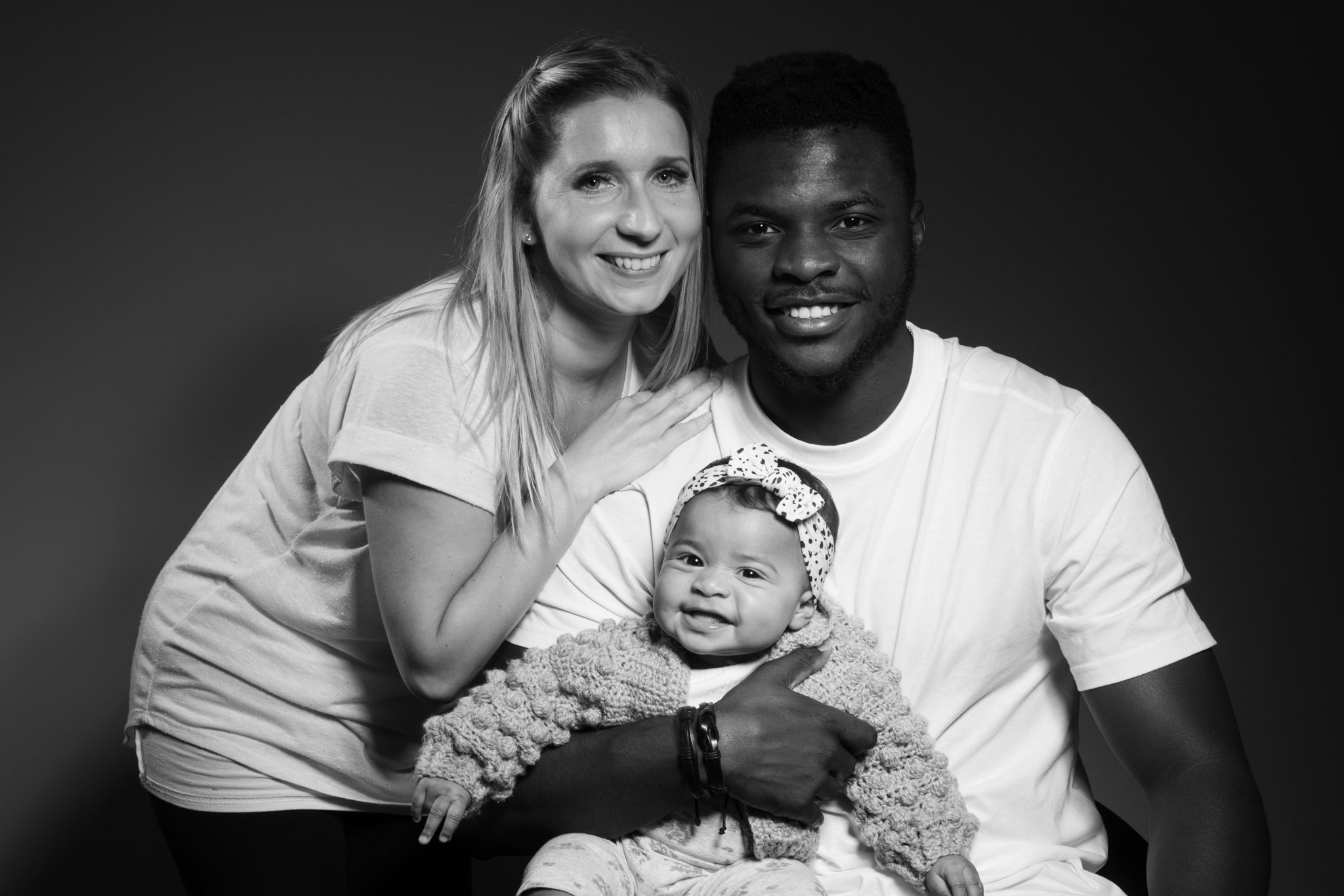 A smiling woman with long hair, a smiling man with short hair, and a young girl with a bow, pose together for a portrait against a dark background.