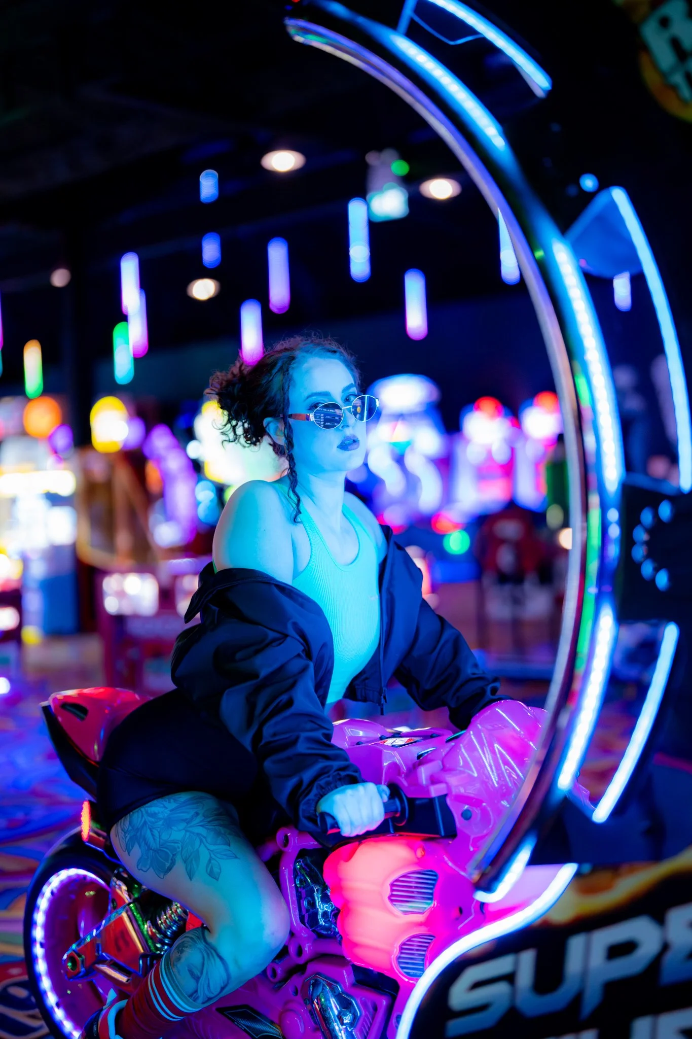 A woman with curly hair, sunglasses, and a blue top riding a pink neon-lit motorcycle arcade game at an amusement park with colorful lights in the background. photographed in Walton, essex in the arcades on the pier. Anne Wills Photography 