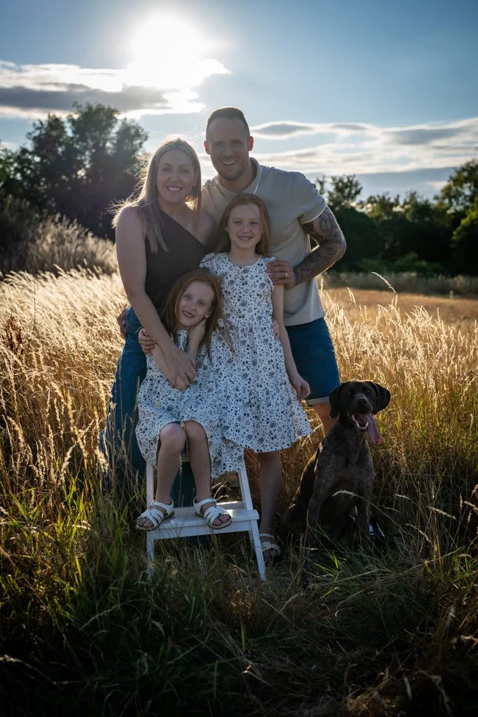 A family of four, two adults and two young girls, standing in a grassy field with a dog, during sunset.