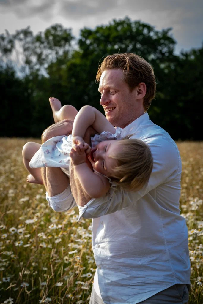 A man holding two young children in a field of daisies, smiling and enjoying a sunny day. Anne Wills Photography. Braintree, Essex