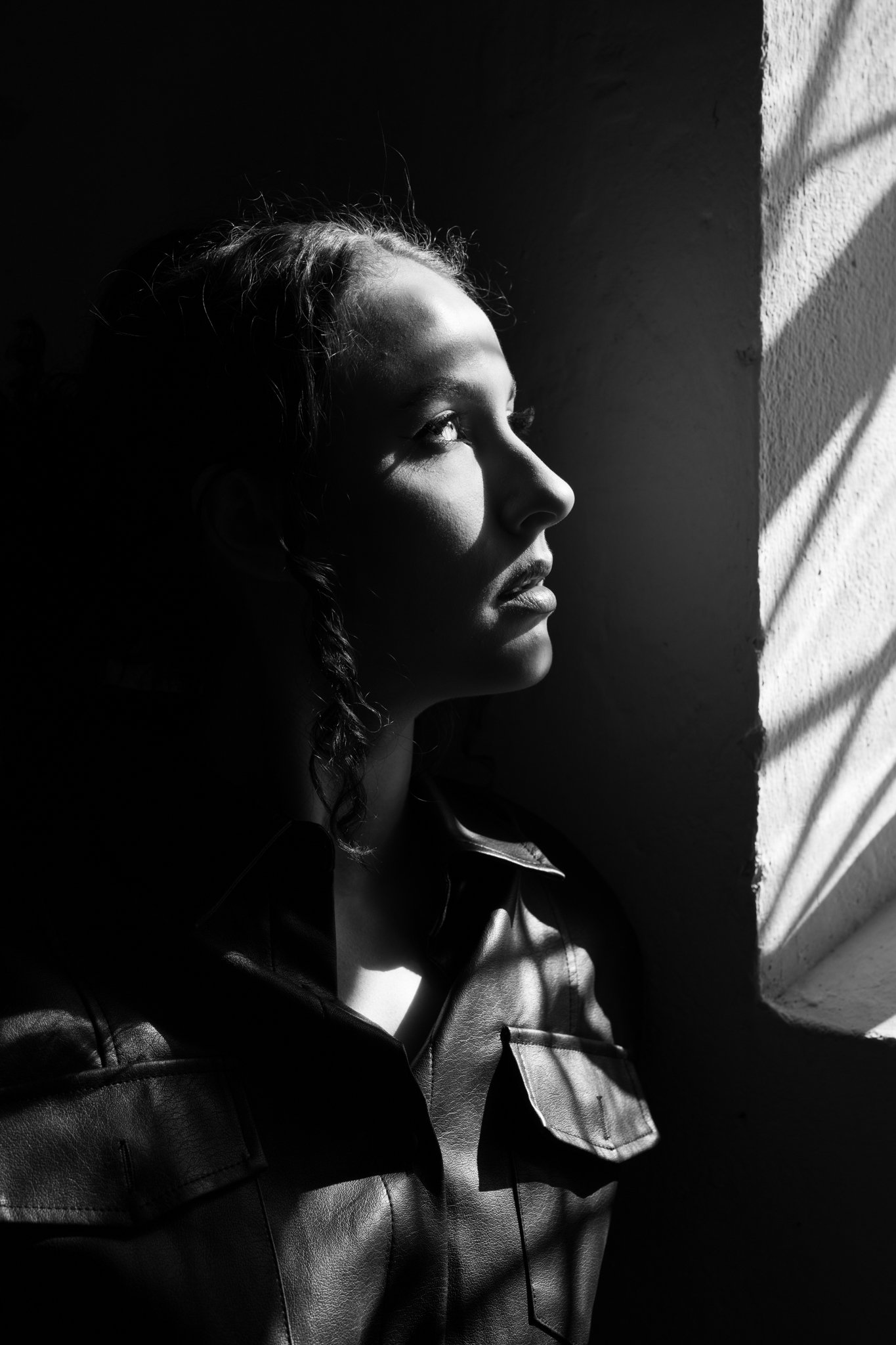 Black and white photo of a woman with curly hair looking towards a wall with sunlight casting shadows on her face.