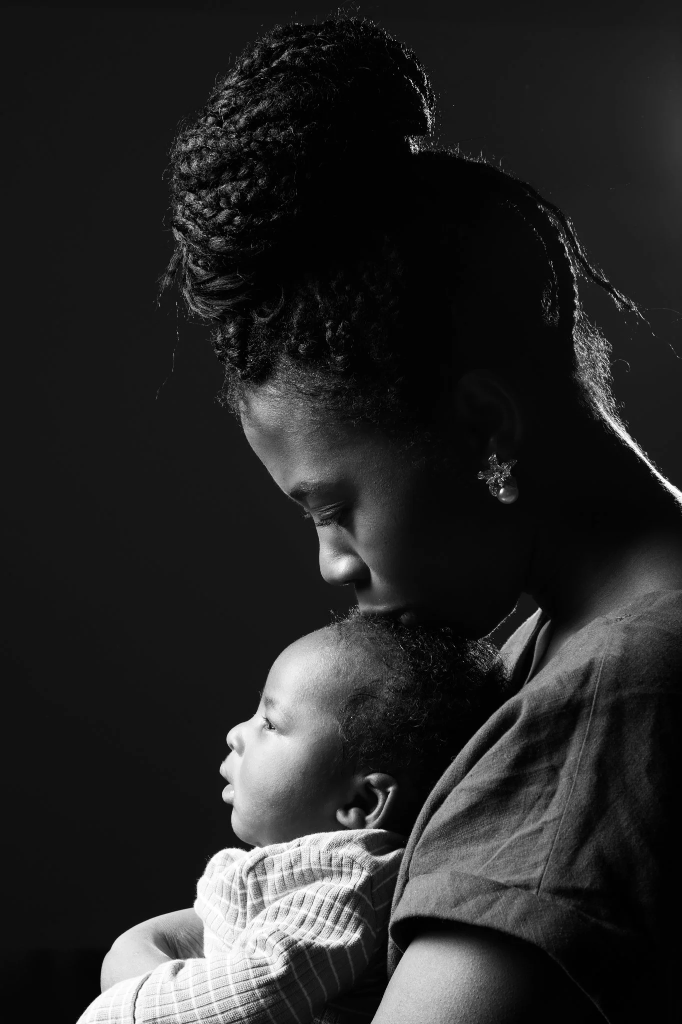A black and white photograph of an African-American woman with her hair styled in an updo, holding a young child in her arms. The woman has a protective and serene expression, wearing an elegant pearl earring, and the child appears to be a toddler wi