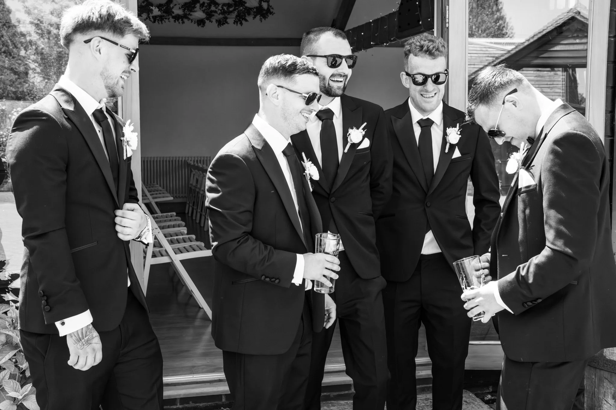 Group of six men dressed in tuxedos and sunglasses, smiling and chatting outdoors at a celebration or wedding. Anne Wills Photography Maidens barn Essex