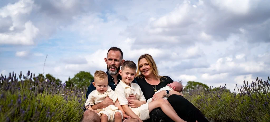 A family of five sitting in a field of lavender, with cloudy sky overhead. The parents are holding their children, including a newborn.
