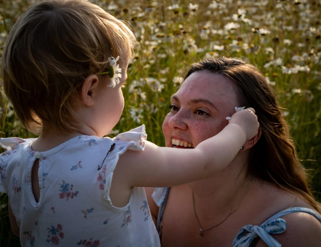 A young girl and a woman, likely her mother, sharing a joyful moment outdoors in a flower-filled field during sunset. Anne Wills Photography. Braintree, Essex