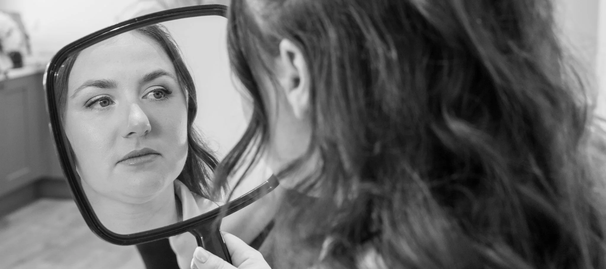 A woman looking at her reflection in a handheld mirror with another person, whose face is not visible, holding the mirror. Anne Wills Photography Maidens barn Essex