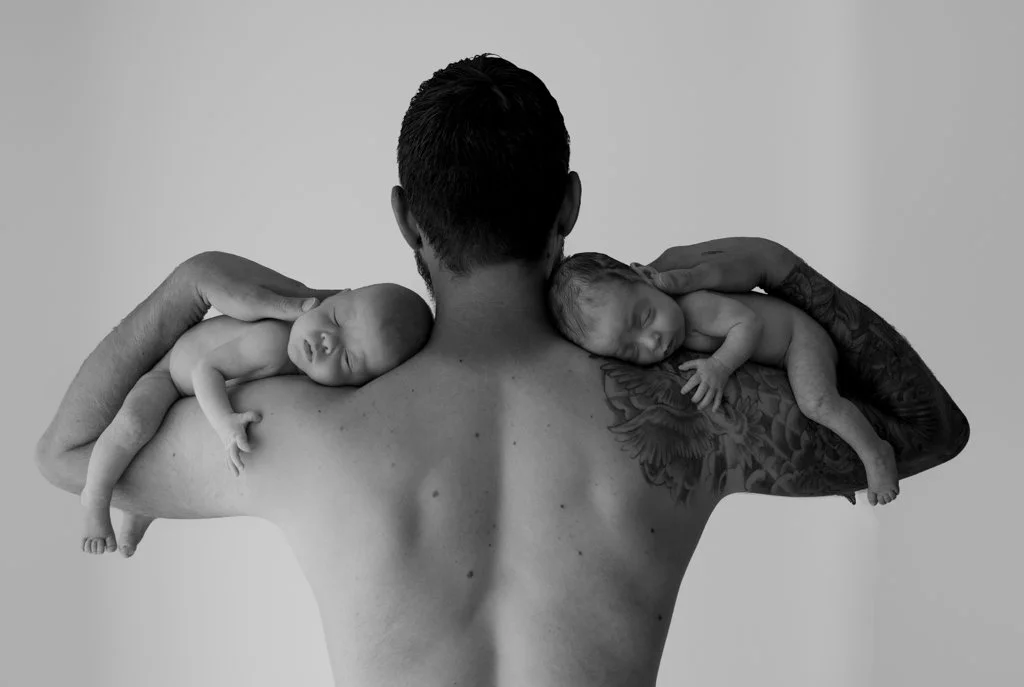 Black and white photo of a tattooed man with short hair, holding two sleeping newborn babies on each shoulder. The background is plain and light-colored.