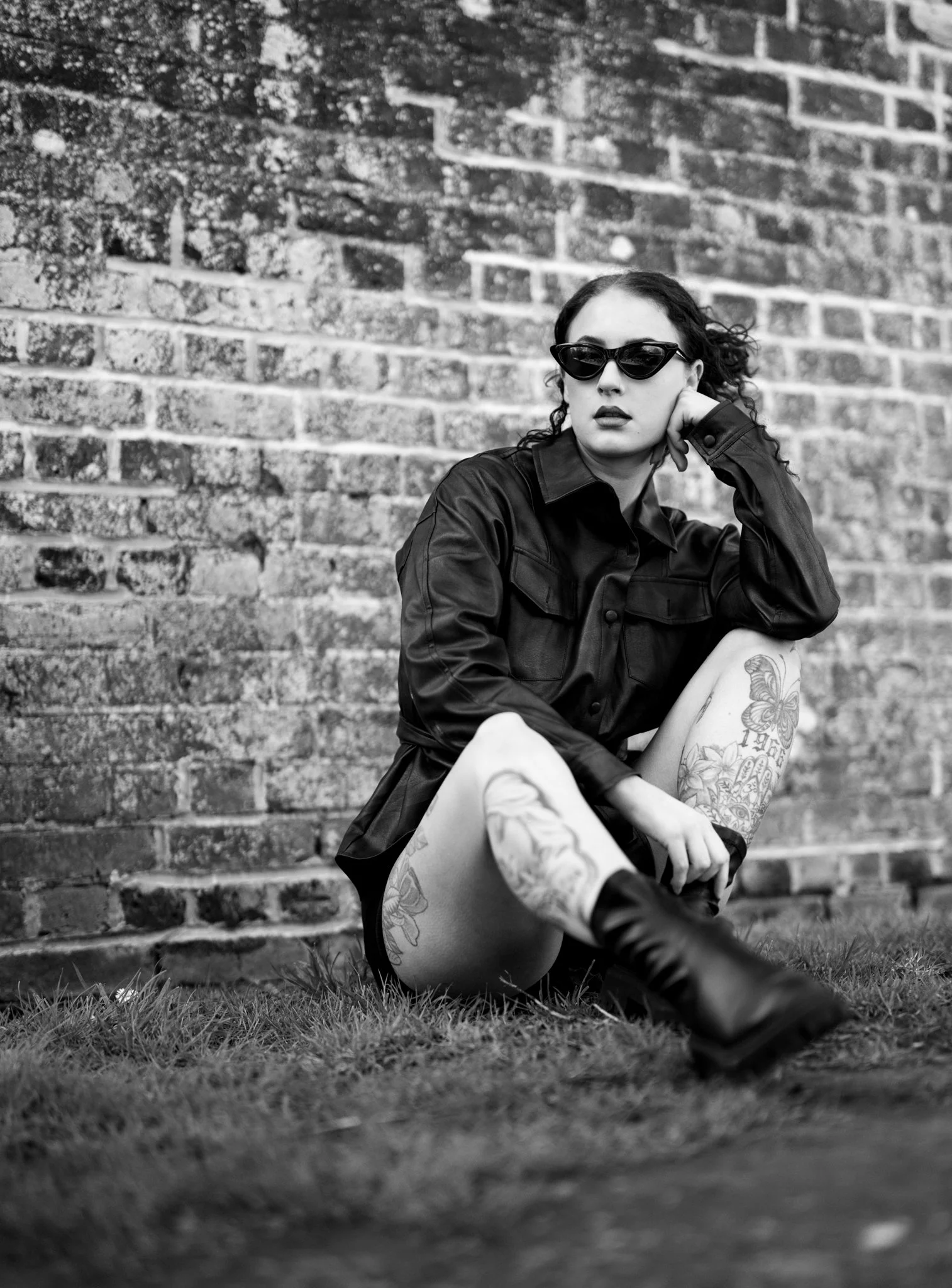 A woman with dark curly hair wearing sunglasses and a leather jacket sitting against a brick wall. She has tattoos on her leg and arm, and she appears to be posing with her hand resting on her face. in earls colne, Essex. Anne Wills Photography 