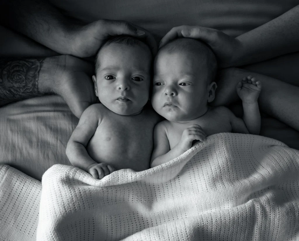 Two infants lying side by side, covered with a blanket, with adult hands gently cradling their heads, in a black and white photograph.
anne wills photography, newborn photography. Braintree, Essex