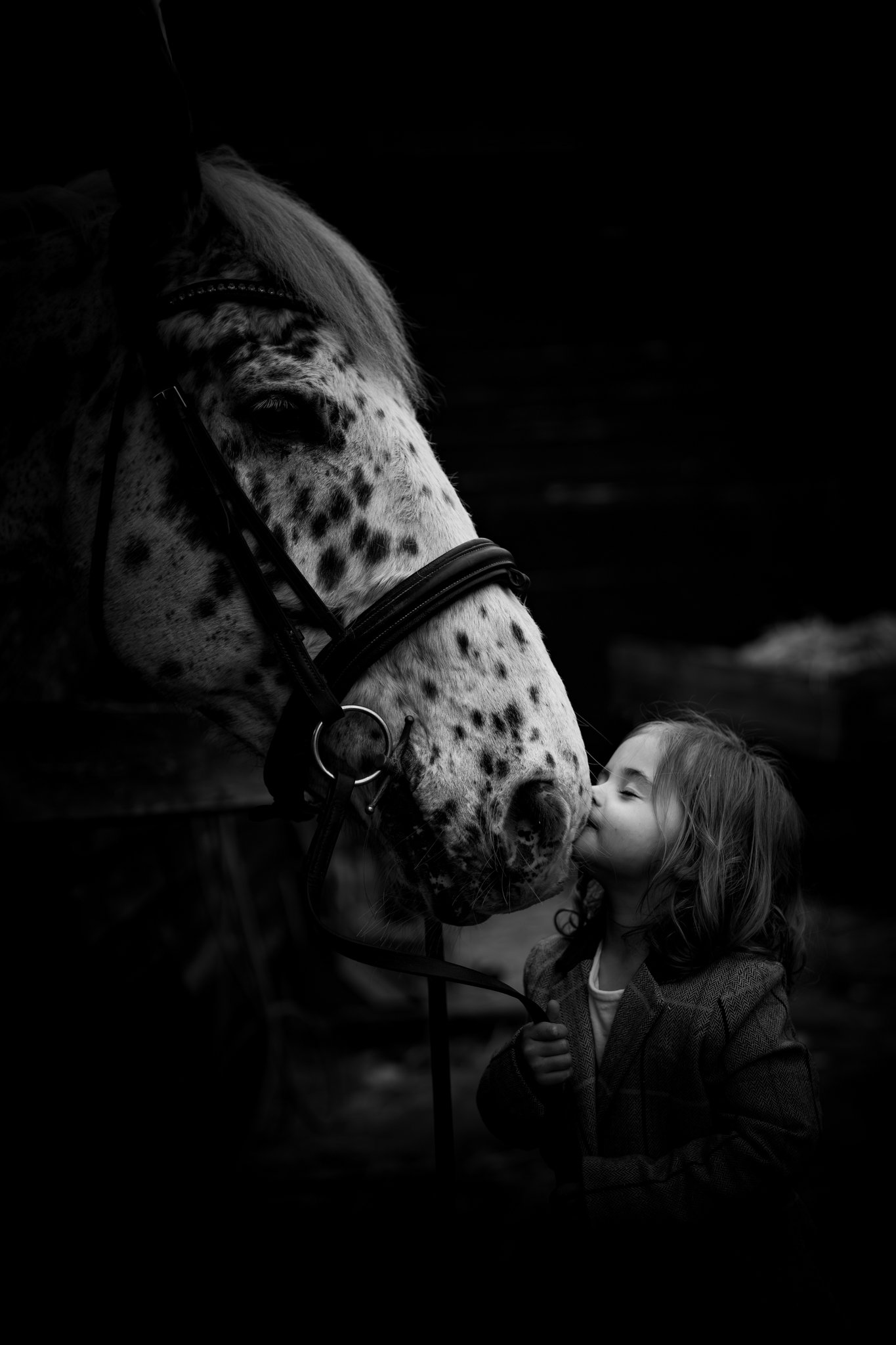 A young girl with curly hair wearing a jacket, standing close to a horse with a spotted coat. The girl is giving the horse a gentle kiss on the nose, and they are outdoors in a dark, possibly cloudy setting.