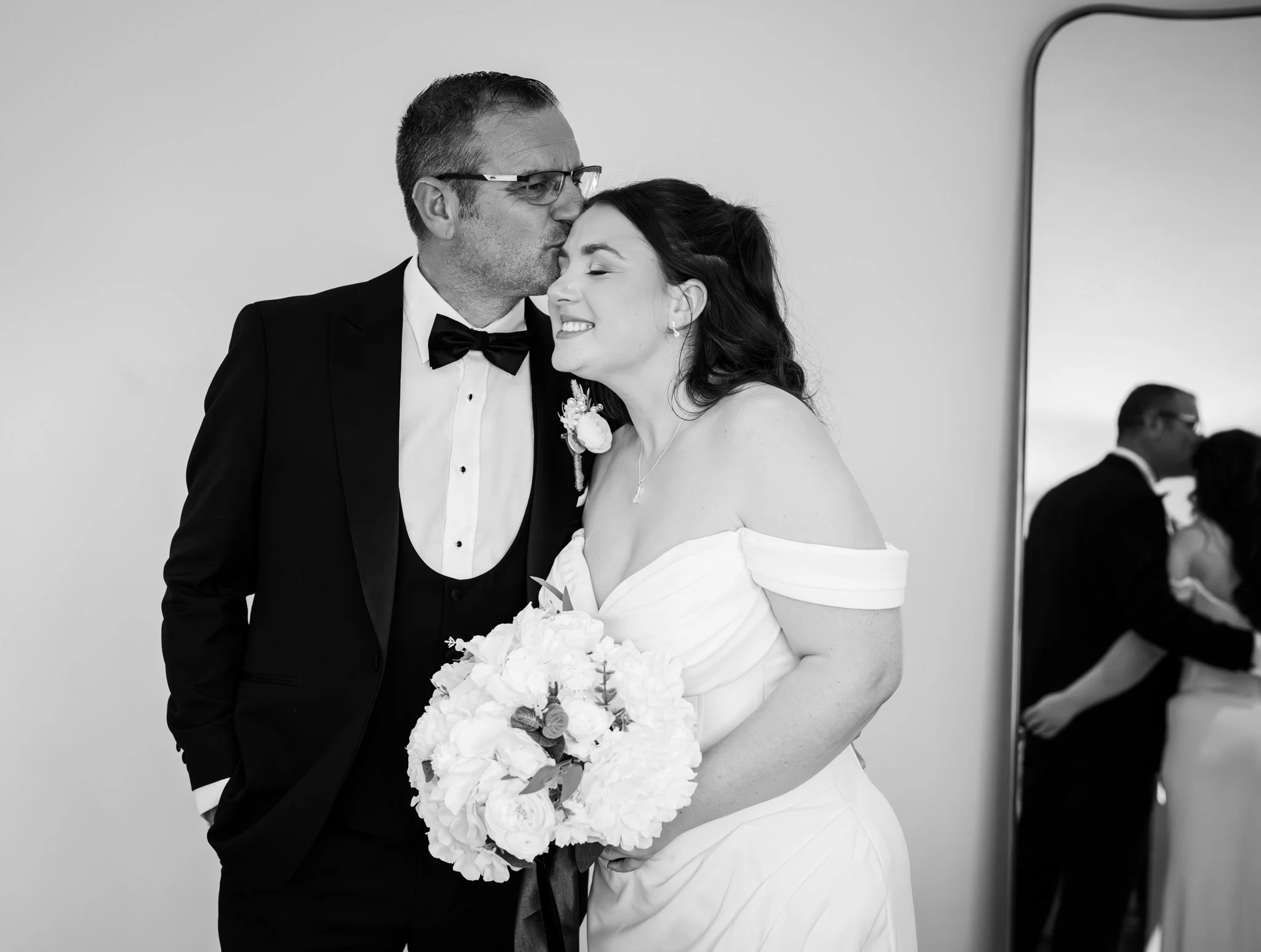 Black and white photo of a groom kissing a bride on her forehead indoors, with a mirror and other people blurred in the background. Anne Wills Photography Maidens barn Essex
