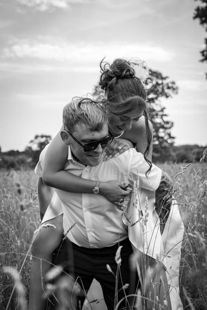 A man wearing sunglasses and a woman with a bun hairstyle and flower hair accessory enjoying a piggyback ride in a field of tall grass, smiling and laughing. Anne Wills Photography. Braintree town hall, Essex