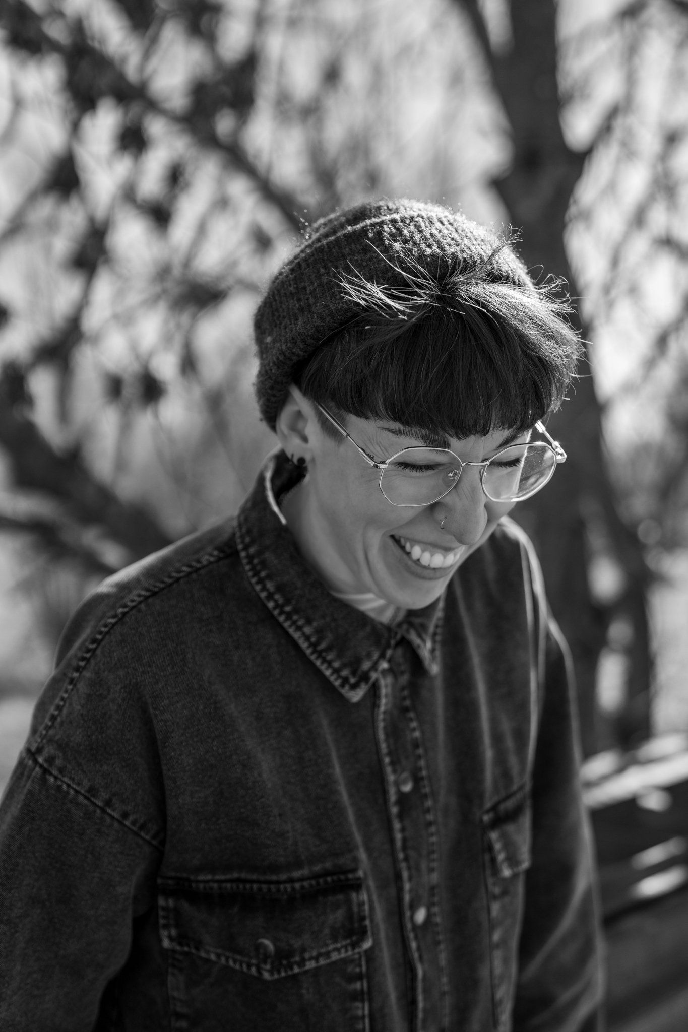 A person smiling outdoors wearing glasses, a knit hat with snow on it, and a denim jacket, with blurred trees in the background.