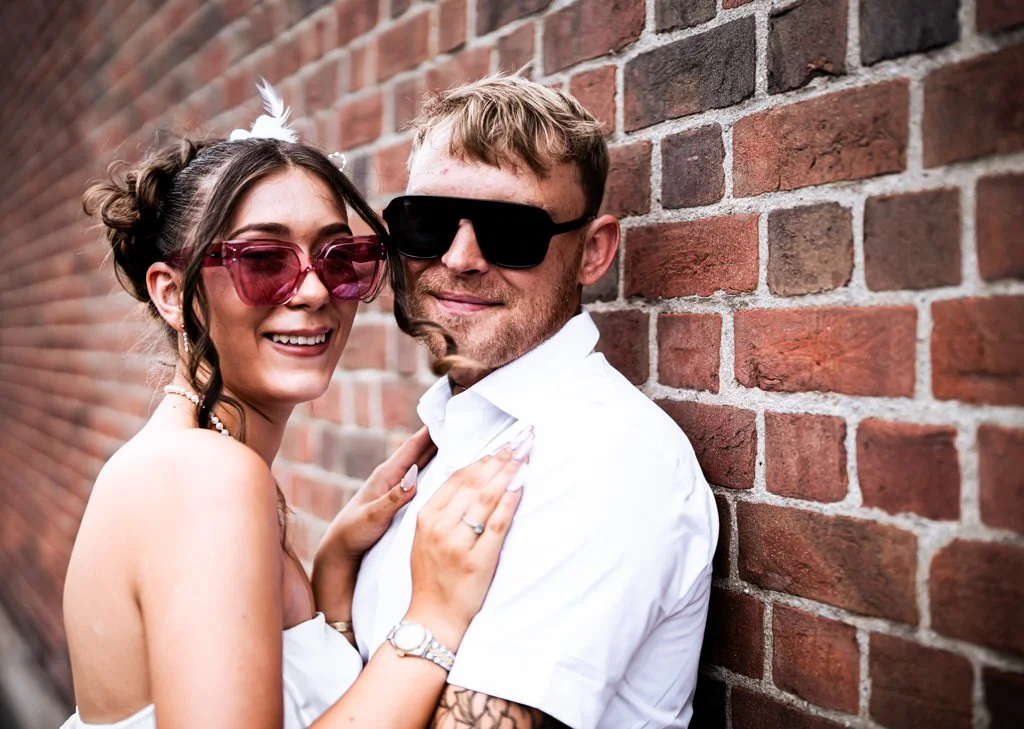 A smiling couple posing close to a brick wall, both wearing sunglasses. The woman has dark hair styled in buns with a white accessory and is holding onto the man's shoulder. The man has light hair, a beard, and is wearing a white shirt. Anne Wills Ph