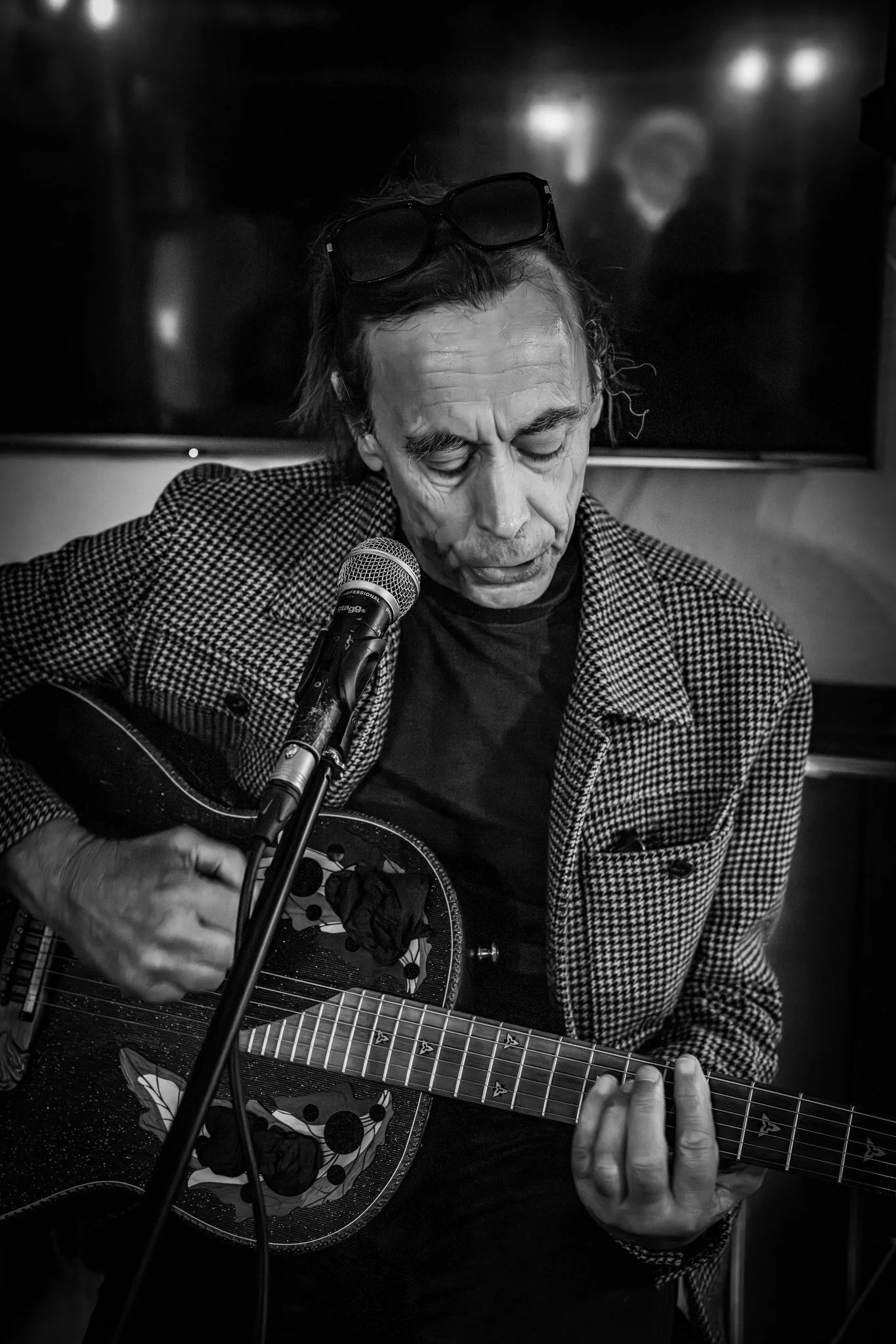 A black and white photo of an elderly man with glasses on his head, playing an acoustic guitar and singing into a microphone. The man has his eyes closed and appears to be emotionally immersed in his music.