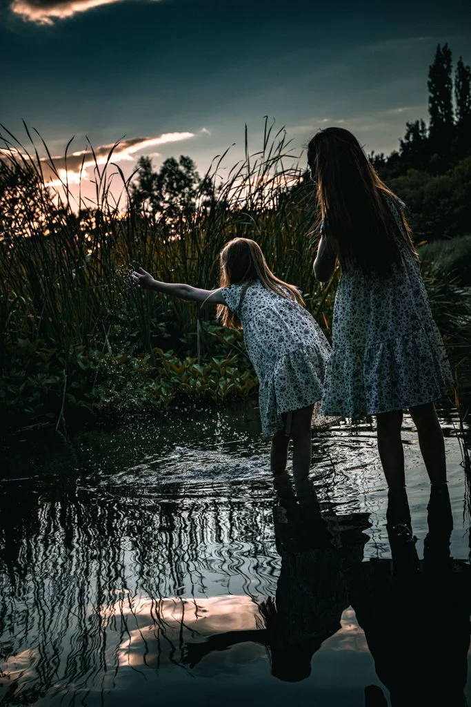 Two girls in floral dresses playing by water at sunset, one splashing water, with trees and a cloudy sky in the background.