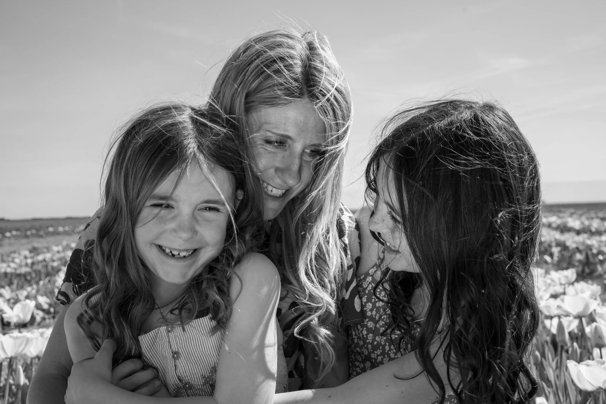 Black and white photo of 3 women, smiling and embracing each other, outside in a field with flowers. hatters farm Essex. Anne Wills Photography 