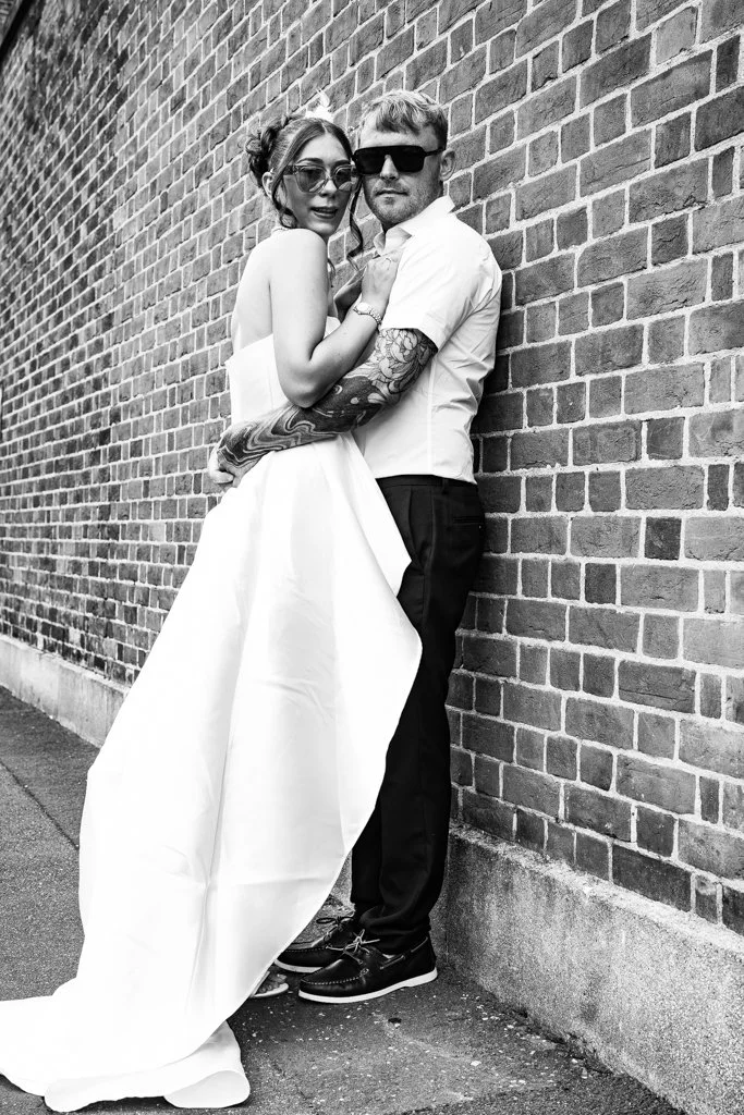 A couple stands close together against a brick wall, posing for a photo. The woman is wearing a strapless white dress and sunglasses, while the man is dressed in a white shirt and black pants, wearing large sunglasses. Both appear stylish and confide