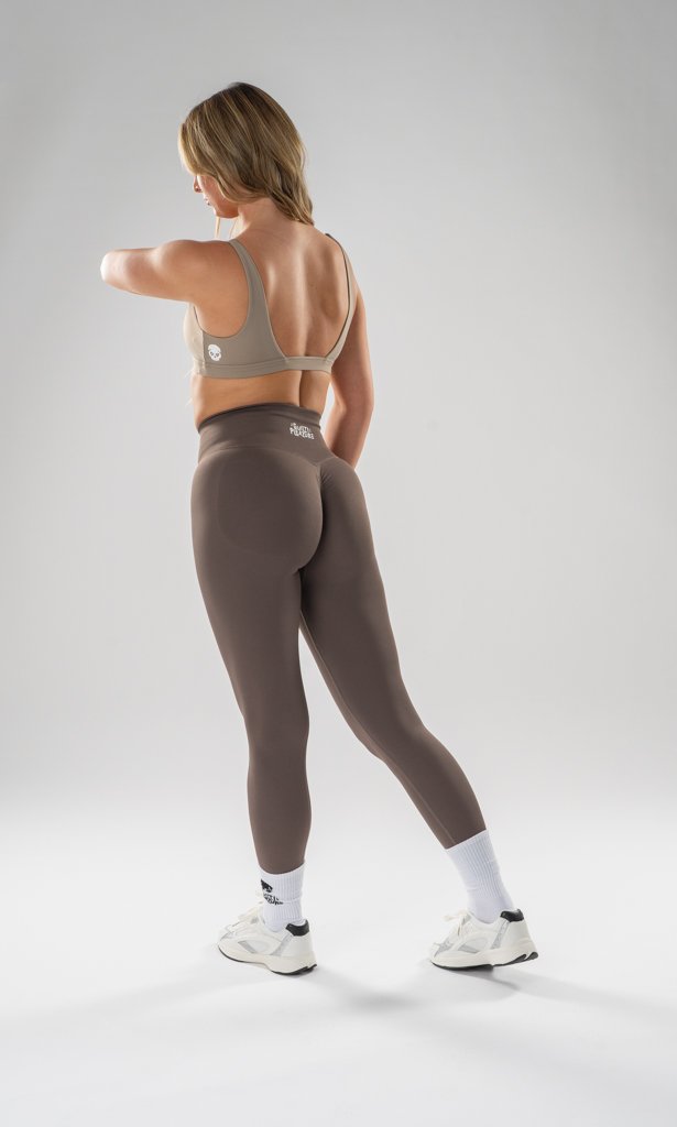A woman with blonde hair standing with her back to the camera, wearing a gray sports bra, taupe leggings, white athletic shoes, and white socks, in a neutral studio setting.