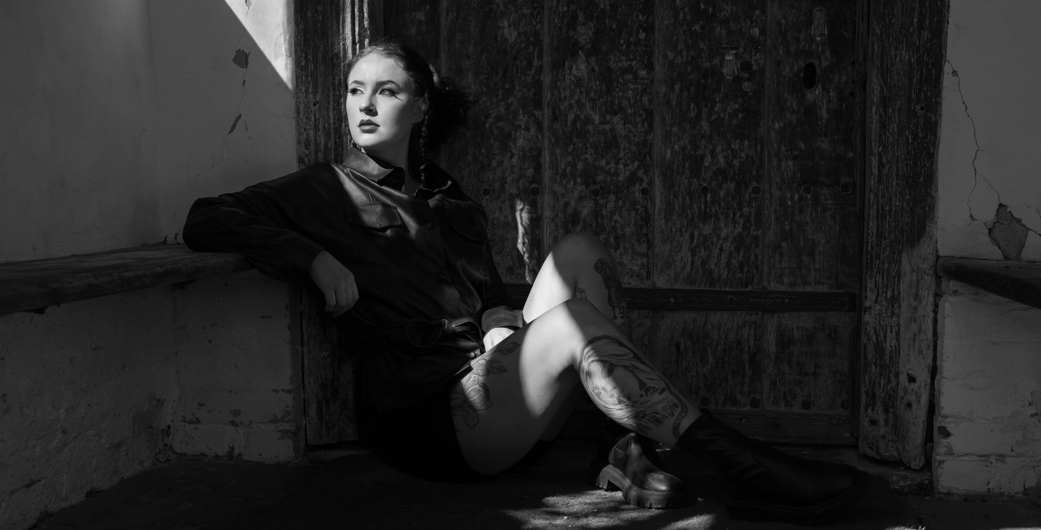 Black and white photo of a woman sitting indoors, leaning against a ledge with her left arm supporting her. She has wavy hair, bold eye makeup, dark lipstick, and tattoos on her right thigh. The setting appears to be an abandoned or rustic space with