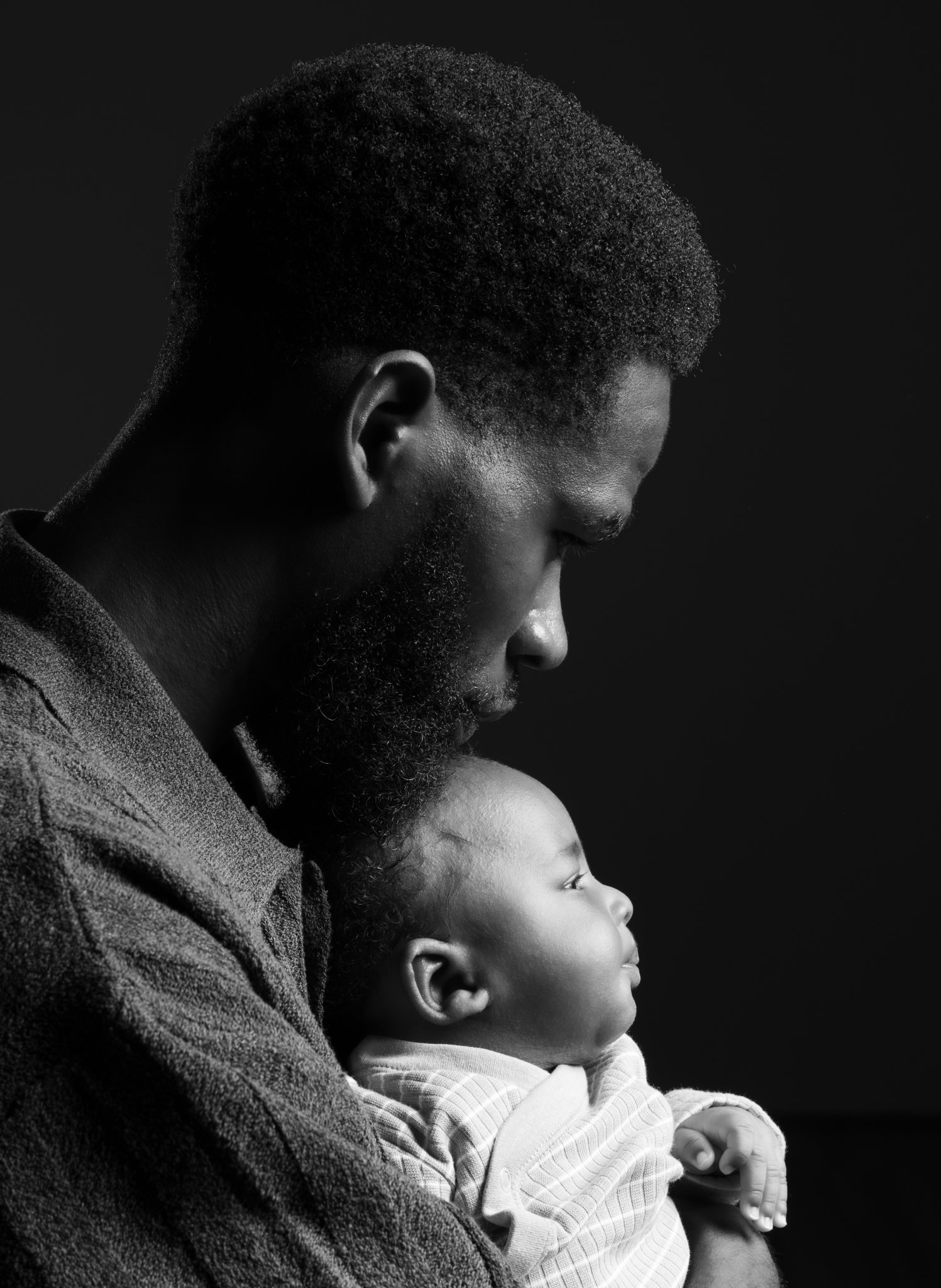 A black and white photograph of a man with curly hair and a beard holding a young child with short hair and a light-colored striped shirt, both facing right against a dark background. Anne Wills Photography Portrait photography in a studio in Braintr