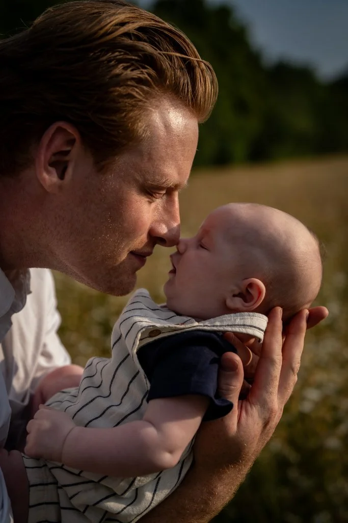 A man with reddish hair holds a baby close, touching noses outdoors with a blurred natural background, creating an intimate moment. Anne Wills Photography. Braintree, Essex