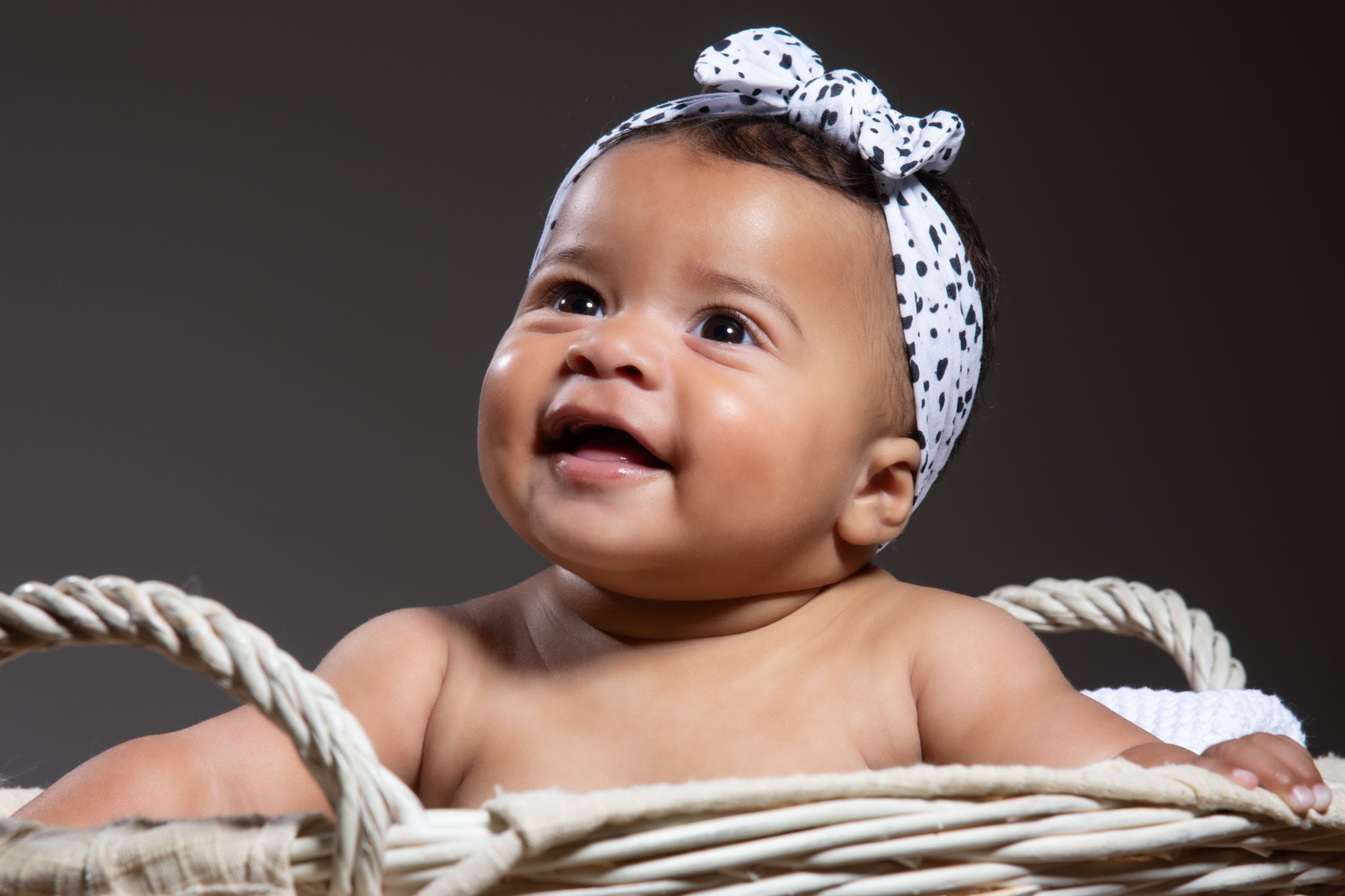 A smiling baby with dark hair wearing a white headband with black spots, sitting in a white woven basket. photographed in Braintree Essex. Anne Wills Photography 