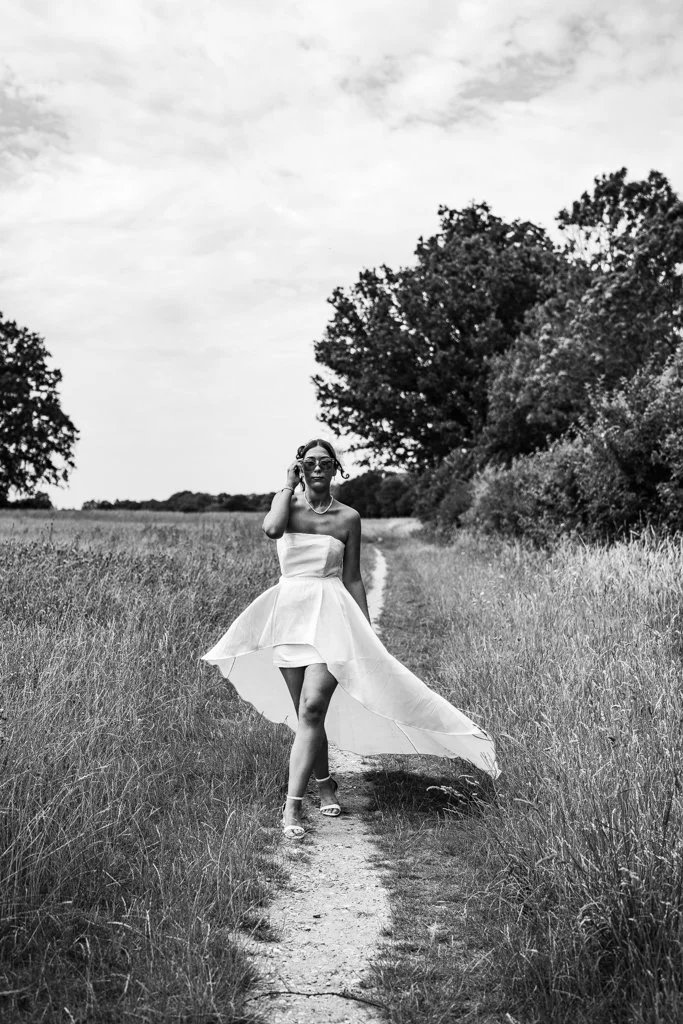 A woman in a strapless dress and sunglasses walking on a dirt path through a grassy field with trees in the background, black and white photo. Anne Wills Photography. Braintree, Essex