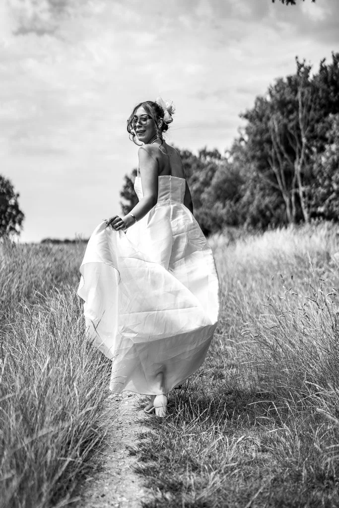 A woman in a wedding dress walking on a grassy path in a natural outdoor setting, smiling and holding the dress.