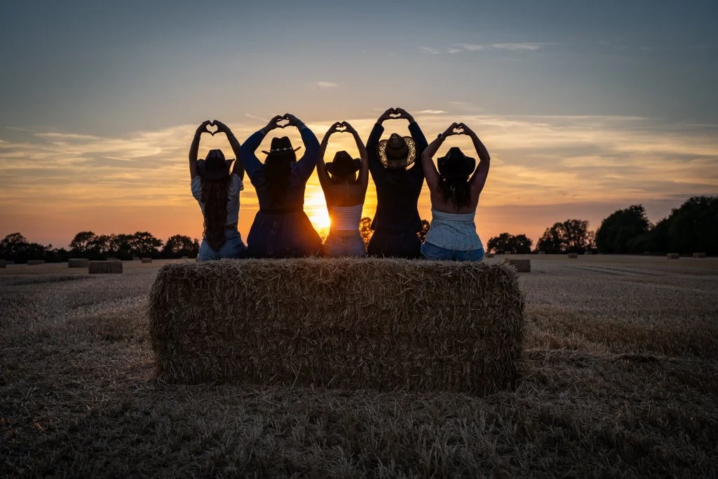Five women sitting on a hay bale in a field at sunset, wearing hats, making heart shapes with their hands above their heads.