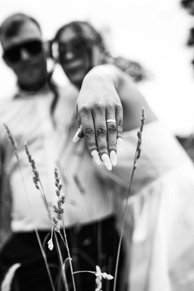 A close-up of a woman showcasing her hand with manicured nails, a ring, and jewelry while the couple stands behind her outdoors in a field, both wearing sunglasses. Anne Wills Photography. Braintree town hall, Essex