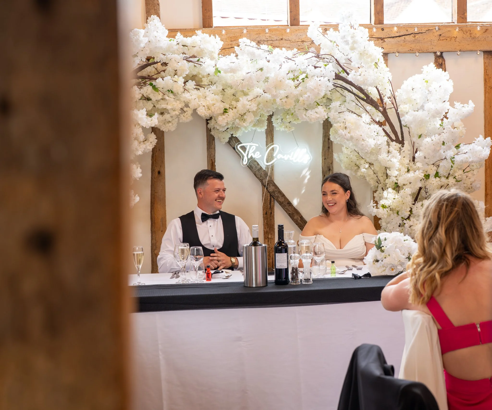 A wedding reception with a bride and groom seated at a table decorated with white flowers and wine bottles, a man and woman smiling and facing each other, under a backdrop of large white floral arrangements and a neon sign that reads "The Covins" in 