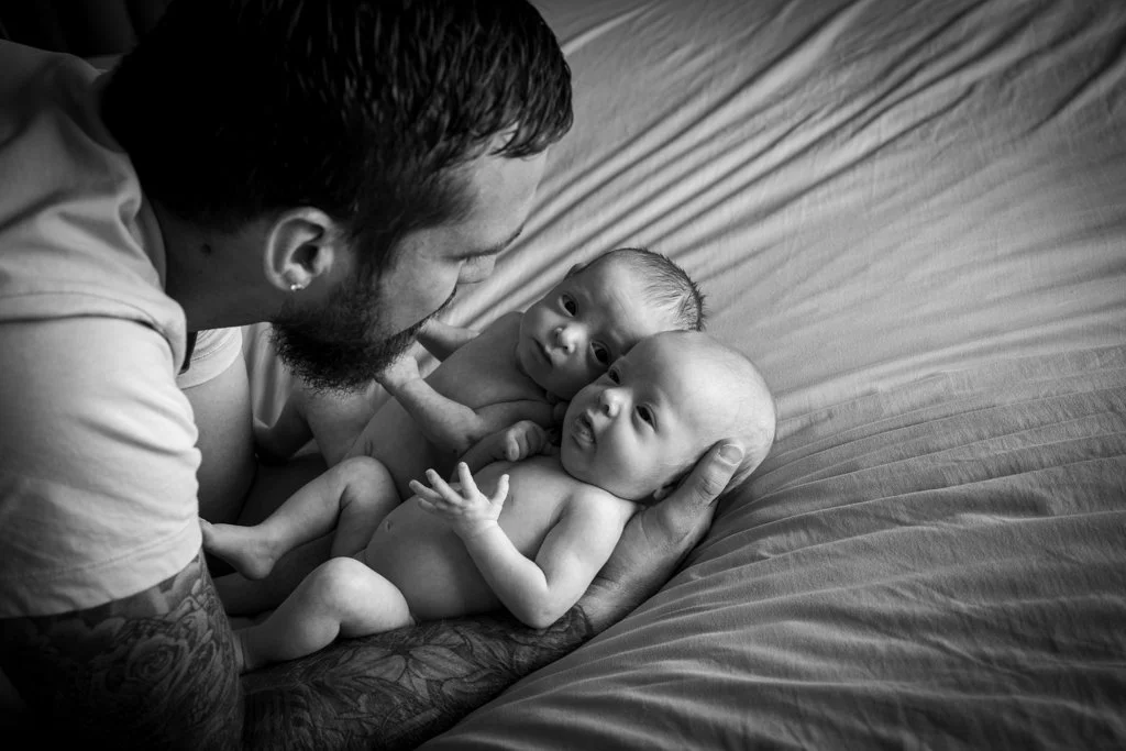 A man with tattoos holding two newborn babies on a bed, looking at them lovingly, in black and white.