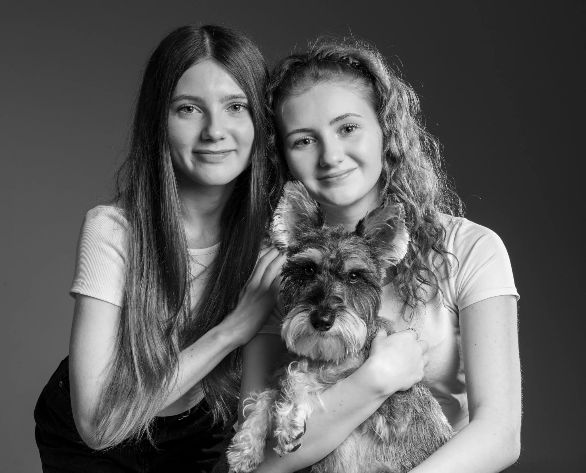 Two young women with long hair, one with straight hair and the other with curly hair, smiling and holding a fluffy dog, all facing the camera against a dark background. photographed in Braintree essex. Anne Wills Photography 